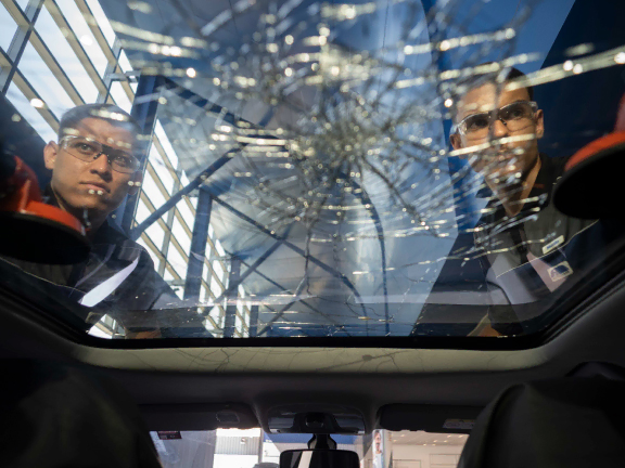 Dois lojistas da Autoglass observando o interior de um carro com teto de vidro estilhaçado, provavelmente após um acidente, em um ambiente interno bem iluminado.