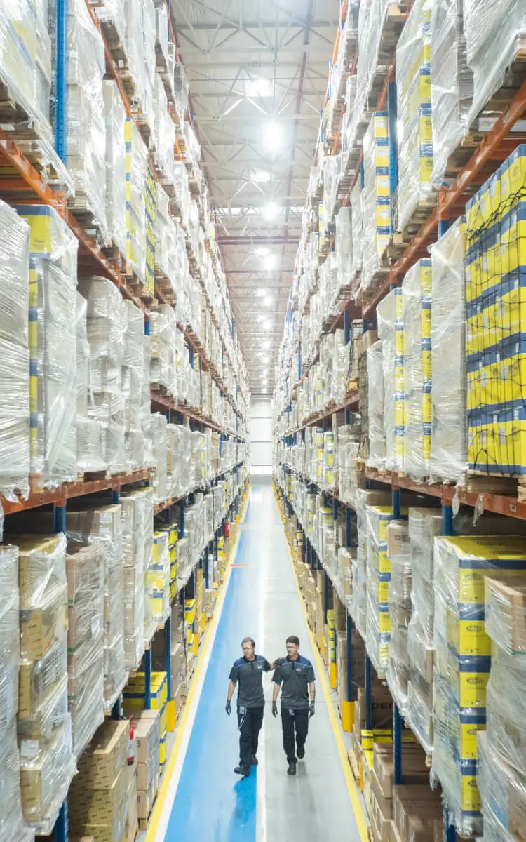 Imagem de dois seguranças patrulhando corredor de armazém com estantes altas cheias de caixas embaladas, mostrando organização e segurança na logística de armazenamento.
